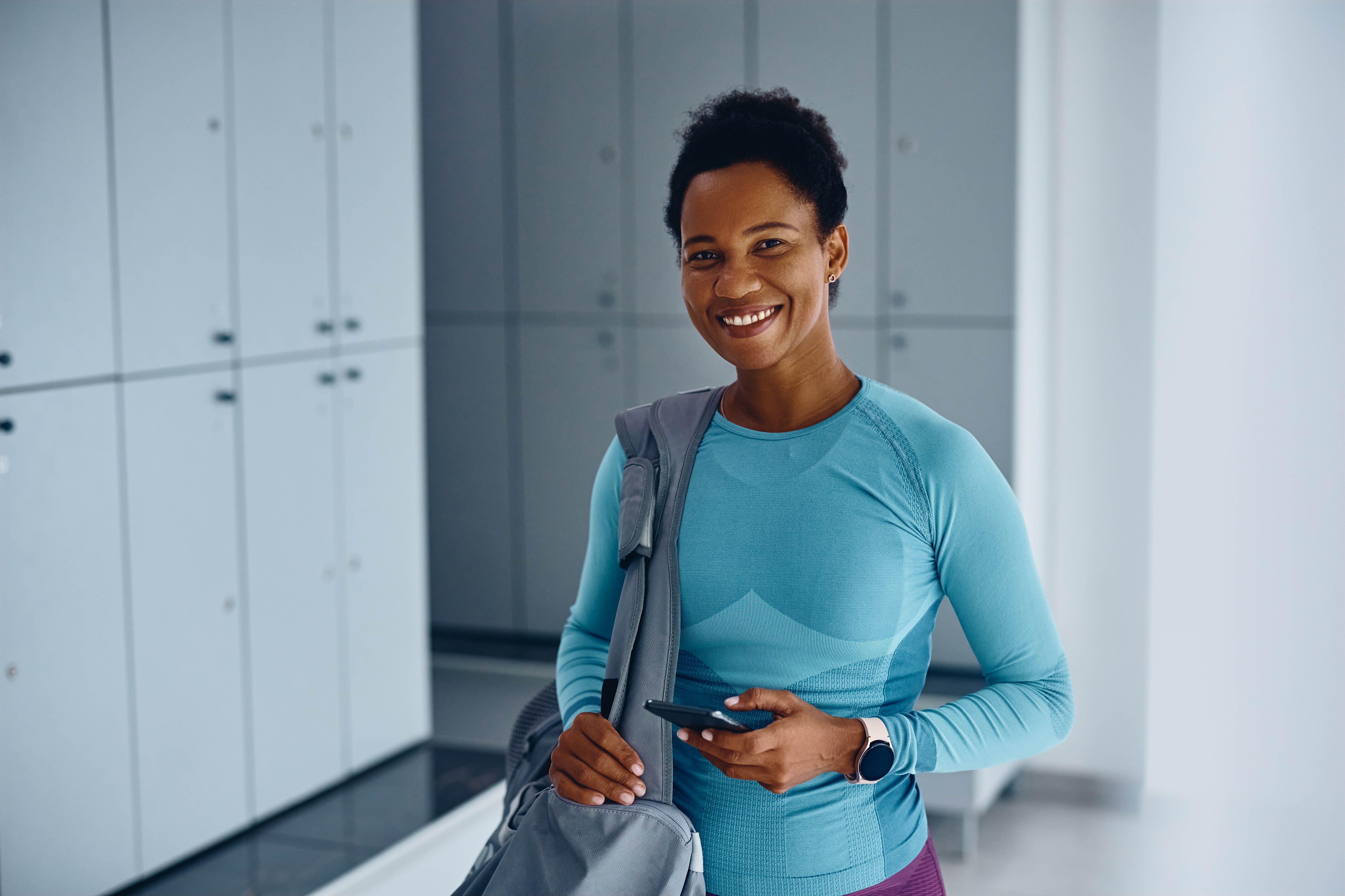 Middle aged woman standing and smiling, with her athletic gear in a bag over her shoulder, standing in a gym locker room Middle aged woman standing and smiling, with her athletic gear in a bag over her shoulder, standing in a gym locker room